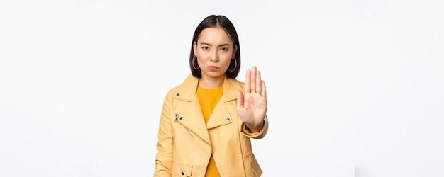 Fille asiatique sérieuse et inquiète étendant le geste d'arrêt de la main de la paume interdire qch interdire debout sur fond de studio blanc