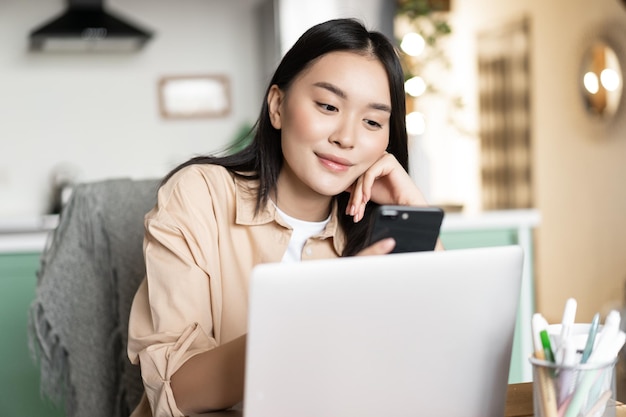 Une fille asiatique souriante étudie en ligne en travaillant sur un ordinateur portable et un smartphone depuis la maison