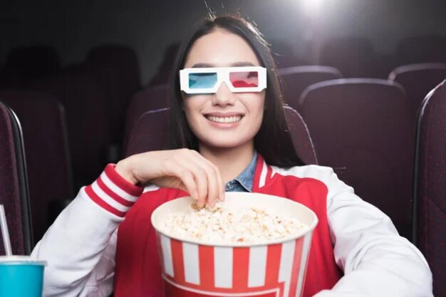 Une fille asiatique souriante avec des lunettes 3D avec un grand panier de pop-corn regardant un film au cinéma