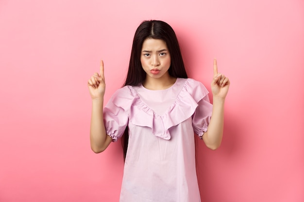 Une fille asiatique triste et maussade regarde sous le front et boude, pointant les doigts vers le haut et se plaignant, regardant jalouse ou bouleversée, debout sur un fond rose.