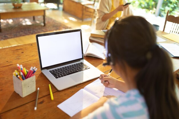 Fille asiatique utilisant un ordinateur portable avec écran vide, écrivant, apprenant en ligne sa mère travaillant en arrière-plan