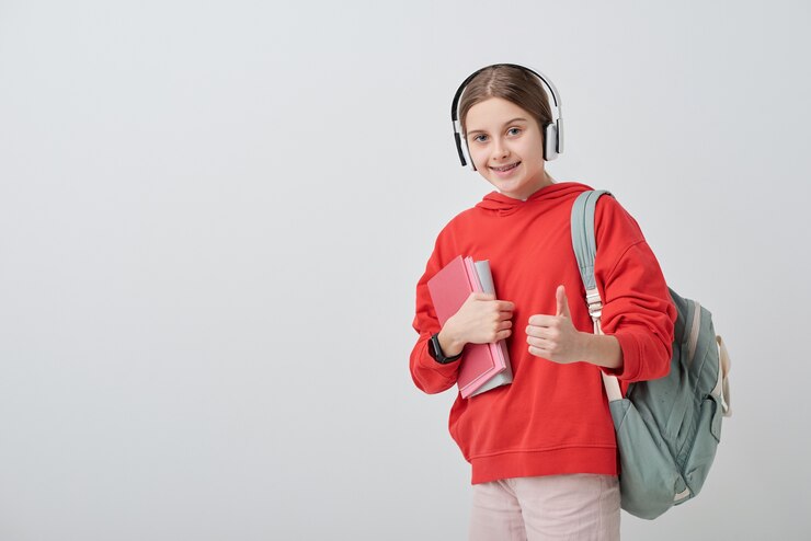Photo fille assez joyeuse avec sac à dos sur le dos et livres à la main montrant le pouce vers le haut tout en écoutant de la musique dans des écouteurs en isolement
