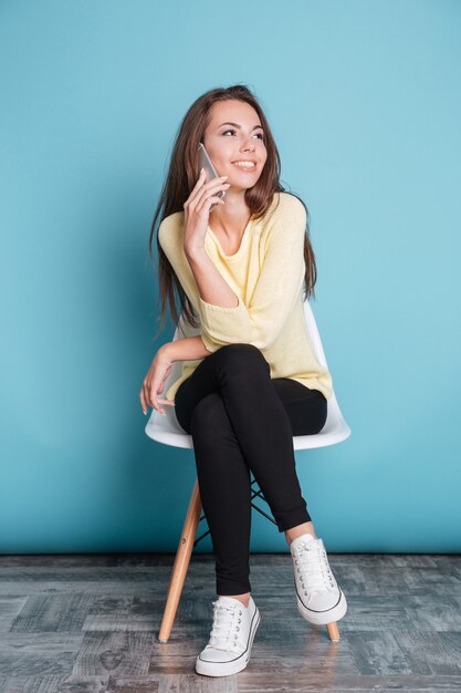 Fille assez souriante parlant sur le smartphone et assise sur la chaise isolée sur fond bleu