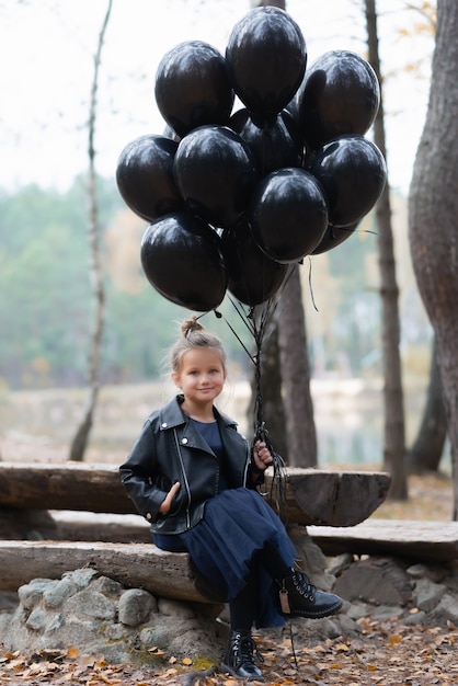 Fille assise sur un banc dans une veste noire et une robe bleue et tenant des ballons noirs