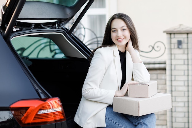 Fille assise dans le coffre de la voiture tenant des boîtes dans ses mains et souriant en regardant la caméra Concept d'achat de biens en ligne et de livraison à domicile