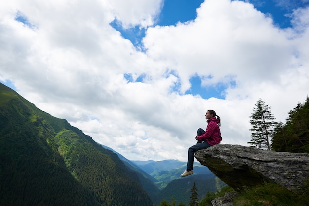 Fille assise sur le rocher, sur fond de montagnes vertes avec des forêts et des nuages au-dessus d'eux à travers lesquels le ciel bleu est visible.