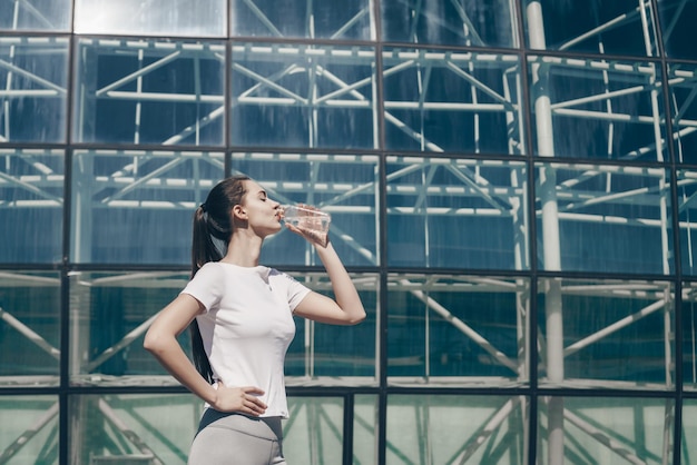 Fille athlétique galbée boit de l'eau propre avant de s'entraîner en plein air