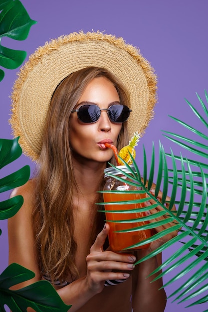 Une fille attirante boit un cocktail de fruits dans des lunettes de soleil et un chapeau de paille tout en se tenant près des plantes tropicales