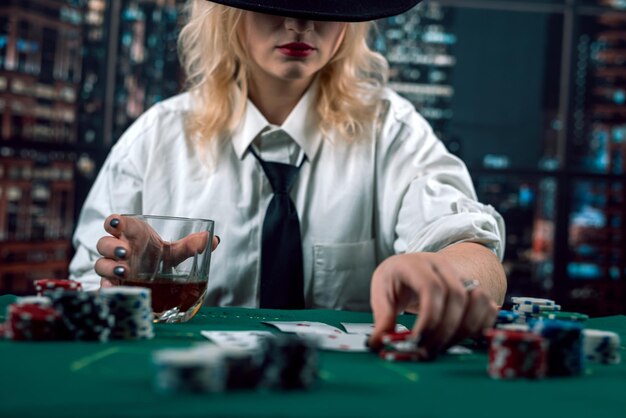 Une fille attrayante en chemise et chapeau buvant du whisky et regardant des cartes de poker au casino.