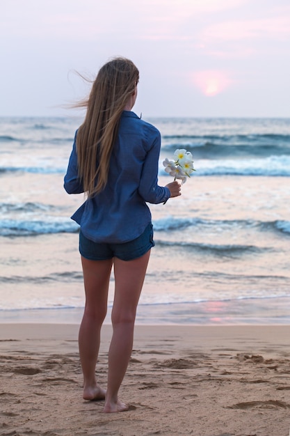 Une fille au bord de l'océan au coucher du soleil, tenant une fleur de plumeria dans sa main