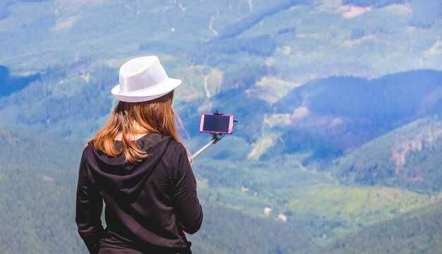 Photo fille au chapeau blanc au sommet de la montagne prend des photos sur téléphone mobile