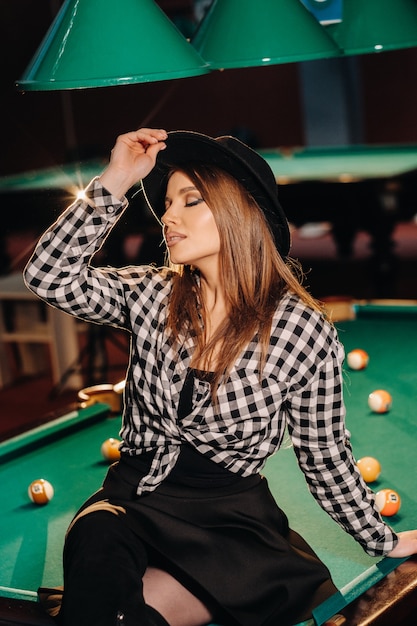 Une fille au chapeau dans un club de billard est assise sur une table de billard. Jouer au billard.