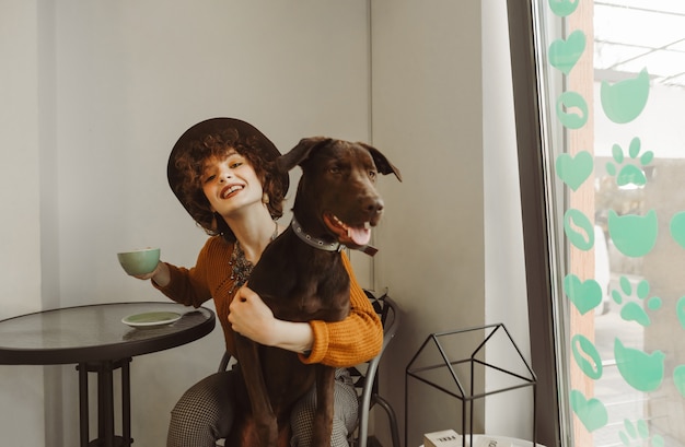 Fille au chapeau est assise dans un café avec une tasse de café et un chien sur ses genoux