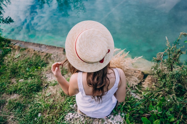 Fille Au Chapeau Est Assise Le Dos Sur Un Fond De Lac Bleu. Fille Un Jour D'été Près De La Source. Fille Dans La Nature.