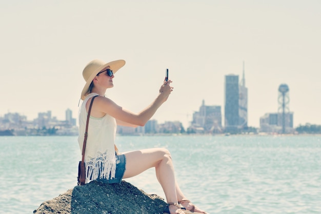 Fille au chapeau fait un selfie assis sur un rocher au bord de la mer