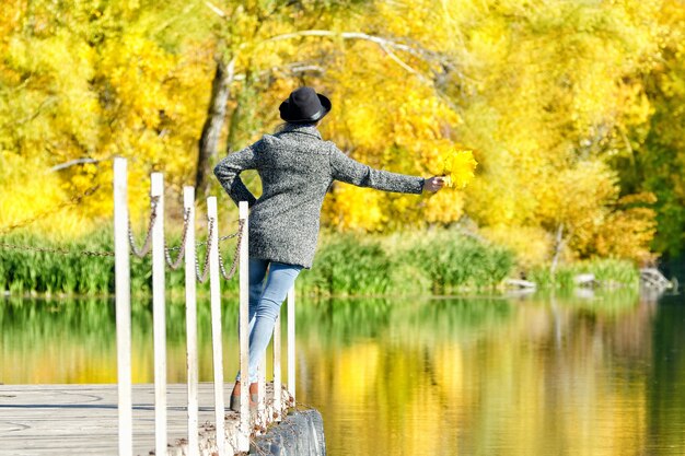 Fille au chapeau avec des feuilles en mains debout sur le quai. Automne, ensoleillé. Vue arrière