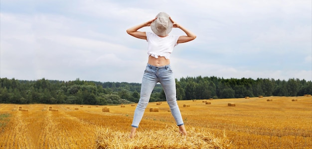 Fille au chapeau de paille se dresse sur une botte de foin sur une balle dans le domaine agricole après la récolte