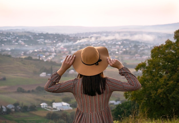 Fille au chapeau par derrière regarde la ville au coucher du soleil