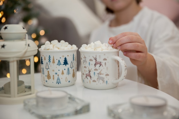 Fille au chapeau de père Noël boit du cacao avec des guimauves au milieu de l'arbre de Noël