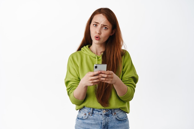 Une fille au gingembre confuse tenant un téléphone et un regard perplexe, un message étrange sur un smartphone, ne peut pas comprendre le fonctionnement de l'application, debout sur fond blanc.