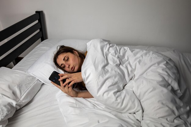 Une fille au lit avec un téléphone sur un lit blanc dans une pièce sombre