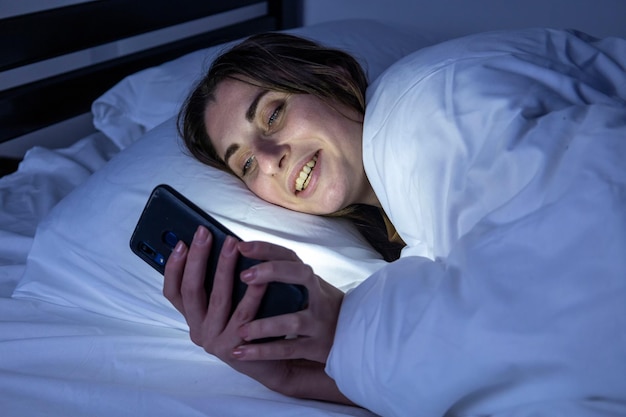 Une fille au lit avec un téléphone sur un lit blanc dans une pièce sombre