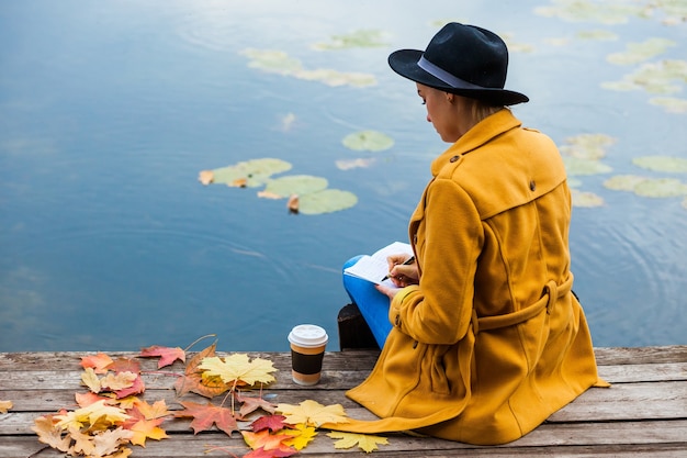 La fille à l'automne est assise au bord de l'eau. L'automne. Feuilles. Rouge jaune. Café