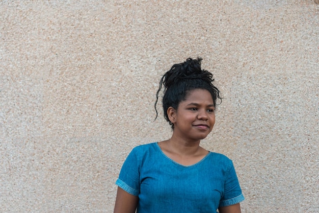 Fille aux cheveux attachés portant une robe debout et regardant sur le côté, avec un mur beige en arrière-plan.