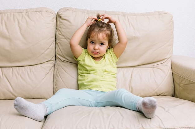 Une fille aux cheveux blonds, assise sur un canapé de couleur crème dans une chemise jaune et un pantalon bleu, touchant sa tête