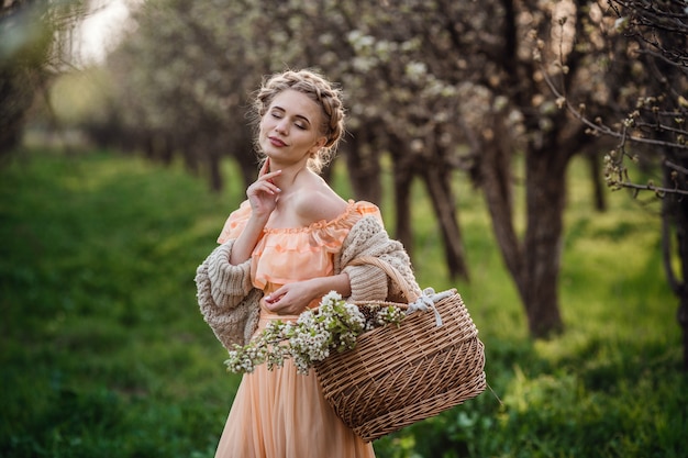 Fille aux cheveux blonds dans une robe légère dans un jardin fleuri. Fille dans une belle robe et pull tricoté bénéficie du coucher du soleil dans un jardin fleuri de poiriers, avec un panier de fleurs