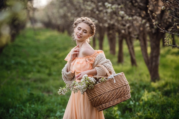 Fille aux cheveux blonds dans une robe légère dans un jardin fleuri. Fille dans une belle robe et pull tricoté bénéficie du coucher du soleil dans un jardin fleuri de poiriers, avec un panier de fleurs