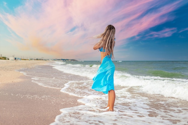 Une fille aux cheveux blonds en maillot de bain bleuté et un châle lumineux se promène le long de la plage