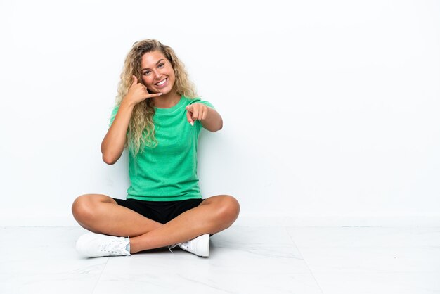 Fille aux cheveux bouclés assis sur le sol faisant un geste de téléphone et pointant vers l'avant