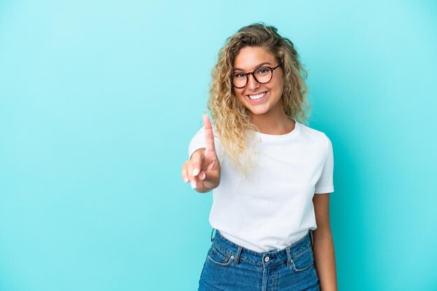 Fille aux cheveux bouclés isolé sur fond bleu montrant et levant un doigt