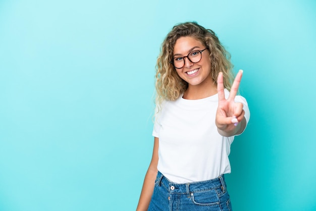 Fille aux cheveux bouclés isolé sur fond bleu souriant et montrant le signe de la victoire