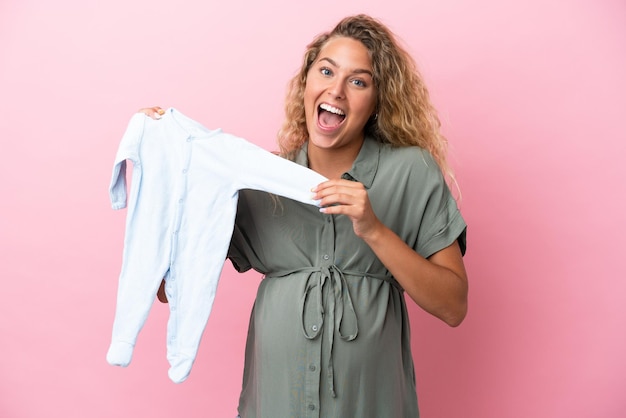 Fille aux cheveux bouclés isolé sur fond rose enceinte et tenant des vêtements de bébé avec une expression surprise