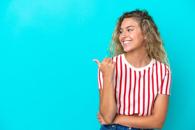 Fille aux cheveux bouclés isolée sur fond bleu pointant sur le côté pour présenter un produit