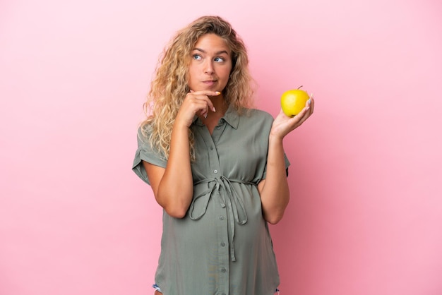 Fille aux cheveux bouclés isolée sur fond rose enceinte et tenant une pomme en pensant