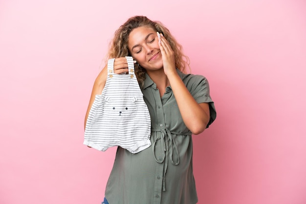 Fille aux cheveux bouclés isolée sur fond rose enceinte et tenant des vêtements de bébé