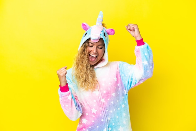 Fille aux cheveux bouclés portant un pyjama licorne isolé sur fond jaune célébrant une victoire