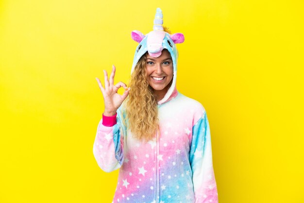 Fille aux cheveux bouclés portant un pyjama licorne isolé sur fond jaune montrant le signe ok avec les doigts