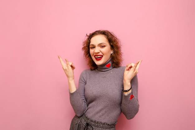 fille aux cheveux bouclés et pull gris se dresse sur fond rose regarde à huis clos avec un sourire sur le visage