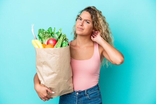 Fille aux cheveux bouclés tenant un sac d'épicerie isolé sur fond vert ayant des doutes