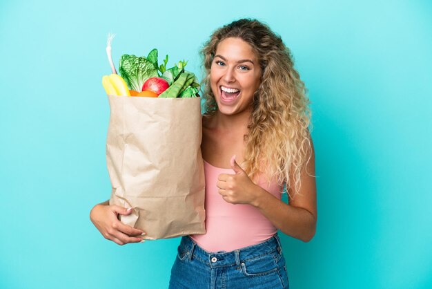 Fille aux cheveux bouclés tenant un sac d'épicerie isolé sur fond vert donnant un geste du pouce levé