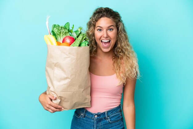 Fille aux cheveux bouclés tenant un sac d'épicerie isolé sur fond vert avec une expression faciale surprise