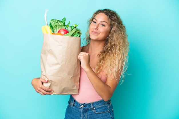 Fille aux cheveux bouclés tenant un sac d'épicerie isolé sur fond vert fier et satisfait de lui-même