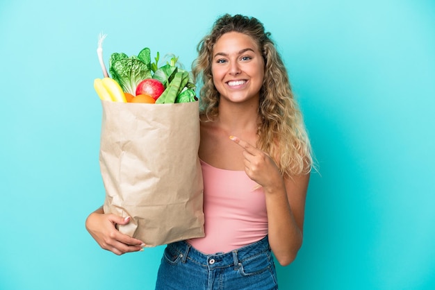 Fille aux cheveux bouclés tenant un sac d'épicerie isolé sur fond vert pointant vers le côté pour présenter un produit