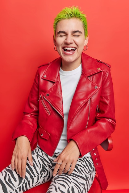 Fille aux cheveux courts teints en vert rit s'amuse d'une apparence extraordinaire vêtue d'une veste en cuir et de leggings pose sur une chaise en rouge