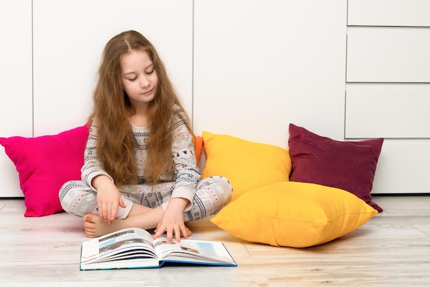 Une fille aux cheveux flottants en pyjama sur le sol parmi des oreillers multicolores lit un livre avec enthousiasme