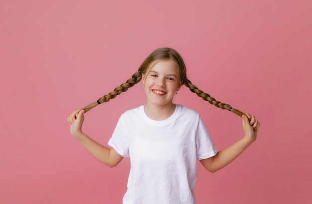 Une fille aux cheveux longs de 1012 ans dans un t-shirt blanc se réjouit et s'accroche aux nattes sur fond rose